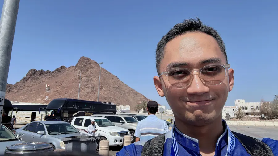 Adryan in glasses and a blue top, squinting in the sun with a rocky mountain behind him